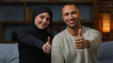 Smiling couple family african american man and arabian muslim woman diverse wife husband sitting on sofa together showing thumbs up like positive recommendation good attitude approve symbol of consent