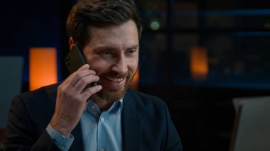 Kafkasyalı bir adam akşam masa başında bilgisayar başında çalışıyor. Cep telefonu, cep telefonu, orta yaşlı iş adamı, 40 'lı yaşlarda, gülümseyen girişimci, akıllı telefonlardan uzak iş konuşmaları yapıyor.