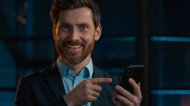 Caucasian bearded businessman happy 30s man worker entrepreneur winner stand in evening office hold cellphone click on mobile phone screen make online order recommend easy pay use gadget smartphone