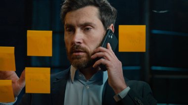 Caucasian businessman marketing assistant middle-aged man multitask talk mobile phone discuss project at office read sticky notes on glass manage business tasks goals analyzing sales strategy research