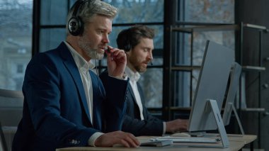 Two men call center service operators work in office talk to client using headset on computer concentrated sales agents sell online answering incoming call provide professional support to customers