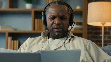 Mature african american man in headphones remote discuss with partner at online conference use laptop at home businessman talk on video call holding documents virtual negotiation concept distance work