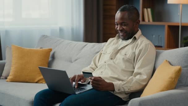 成熟した笑顔アフリカ系アメリカ人男性が自宅でソファの上にラップトップを使用してクレジットカードでオンラインで支払う幸せな男性顧客買い物客は 電子銀行サービスコンピュータアプリケーションを使用してインターネット上で購入のために支払う — ストック動画