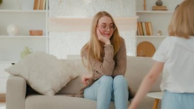 Gülümseyen anne bebek bakıcısı kanepeye otur ve anaokulu kızına bak kanepe etrafında koşuşturuyor mutlu aile anneliği haftasonu evde gürültülü küçük kız çocuk oyunu oynuyor oturma odasında.