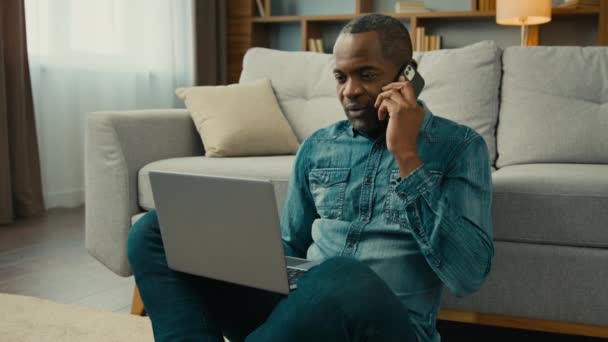 成熟したアフリカ系アメリカ人ビジネスマン携帯電話で話すフリーランスは リモートリモートリビングルームで自宅でノートパソコンを使用してビジネス契約を議論するクライアントに携帯電話の話によるインターネット上での注文を行う — ストック動画