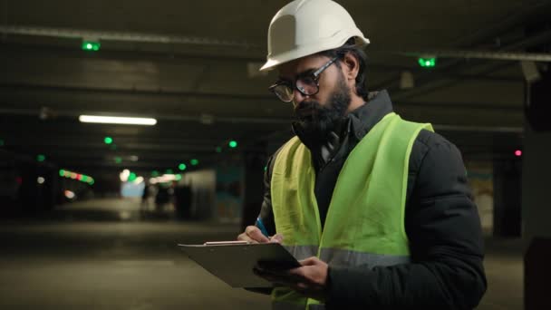 ハード帽子ヘルメットのインドの技術者請負業者の整備士は ハード帽子のチェックで地下の監督アラビア人労働者製造技術者をチェッククリップボードで駐車場を書きます検査 — ストック動画