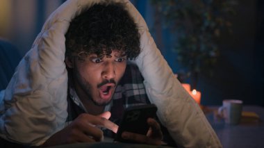 Şok olmuş Hintli adam Arap erkek şaşkınlık haberi okuyor sosyal medyada gece evde battaniyenin altında battaniye altında akıllı telefon mesajıyla şaşkına döndü.