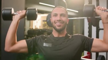 Gülümseyen Afrikalı Amerikalı sporcu spor salonunda spor aletleri yapan vücut geliştirici ağır halterler kaldıran ağırlık kaldıran erkek sporcu omuz kaslarını çalıştırıyor.