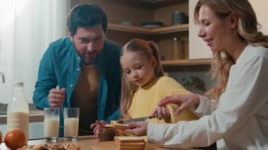 Mutfakta birlikte yemek pişiren Kafkas ailesi küçük kız çocuğuna ekmek üzerine çikolata ezmesi hazırlamasını öğretmeye yardım ediyor.