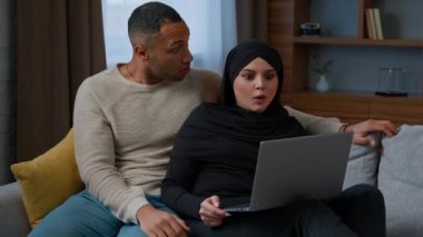 Heyecanlı çift Afrikalı Amerikalı erkek tesettürlü Arap Müslüman kadınla dizüstü bilgisayar kullanıyor. Şaşırtıcı çok ırklı koca konuşması evde internetten alışveriş yap piyango kazan sürpriz zafer kazan