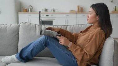 Yan bakışlı Asyalı kadın Kore 'li Japon iş kadını evinde kanepede yatıyor dizüstü bilgisayarla yazışıyor internette sohbet ediyor arama verileri okuyor sosyal medyayı okuyor karantinada internet konforlu serbest çalışmalar yapıyor.