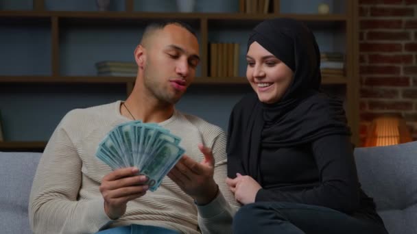 多様なカップル幸せな豊かなアフリカ系アメリカ人男性イスラム教徒女性でヒジャーブカウントお金ドル結婚配偶者は通貨を保持します現金家族の予算ビジネス収益財政は金銭賞金融富を獲得 — ストック動画
