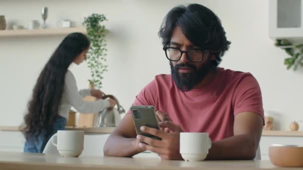 ひげを生やしたイスラム教徒インドのアラビア人男性リモート家庭用キッチンから働く携帯電話アプリの携帯電話をオンラインでチャットを使用しています 夫の背景のための民族の女性の主婦料理朝のコーヒー朝食 — ストック動画