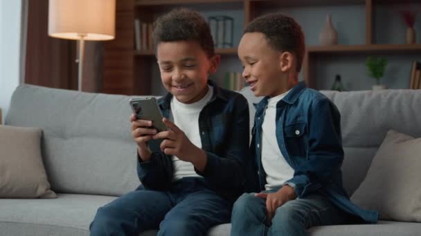 两个快乐的笑着的非裔美国男孩兄弟在家里用智能手机浏览没有父母控制的卡通片 — 图库视频影像