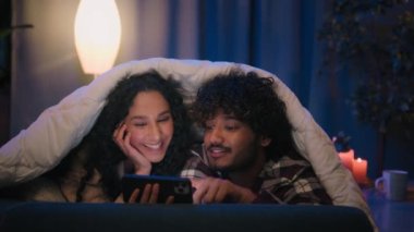 Gece evde cep telefonu kullanan çok ırklı bir çift akşam üstü battaniye altında battaniye altında battaniye altında televizyon karşısında internetteki haber şokuyla şaşkına döndü.