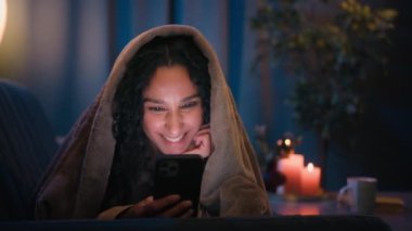 Latin tebessüm eden kız Latin kadın Hintli kadın gece vakti karanlık ev odası örtüsü altında battaniye örtüsü altında cep telefonu bağımlısı akıllı telefon sosyal medya uygulaması
