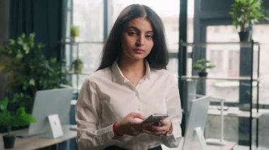 Ofiste cep telefonu kullanan mutlu çok ırklı kadın kameraya gülümseyen Hintli iş kadını Arap kadın yönetici kadın internet sörfçüsü internette geziniyor haber tebessümlerini kontrol ediyor.