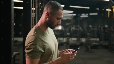 Afrika kökenli Amerikalı sporcu cep telefonlu erkek fitness eğitmeni spor salonunda spor yaptıktan sonra rahatlıyor akıllı telefon teknolojisini tarıyor internet mesajlarına bakıyor kameraya bakıyor