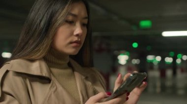 Ciddi Asyalı kadın Çinli Japon kadın etnik bayan müşteri şoförü yeraltı otoparkında cep telefonu uygulaması yazarak internet üzerinden cep telefonu sohbeti yapıyor.