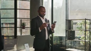 Afro-Amerikalı olgun bir adam, güneşli bir ofiste cep telefonunu tutuyor. Erkek işadamı internet üzerinden alışveriş yapıyor. E-bankacılık uygulaması internet üzerinden ödeme yapıyor.