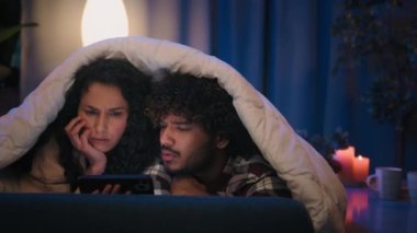 Çok ırklı üzgün çift ve kadın, gece evde hayal kırıklığına uğramış stresi şok etti. Kötü haber cep telefonu, akşam kanepede battaniyeli battaniye altında arıza yaptı.