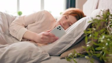 Mutlu, huzurlu, güzel, güler yüzlü, etkili kız blogcu, bağımlı kadın sabah uykusunda rahat bir yatakta, akıllı telefonlu, cep telefonlu, internetteki sosyal medya uygulaması ile sohbet ederek uyanıyor.