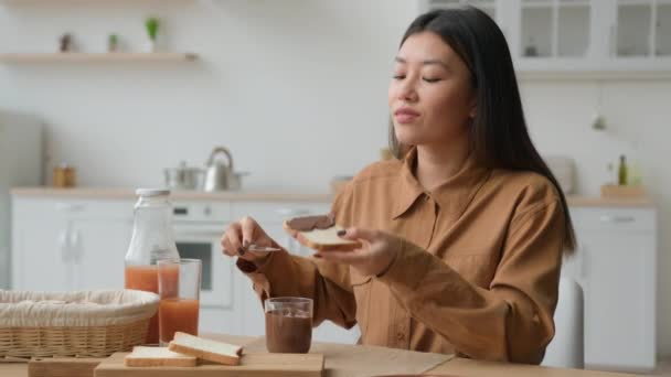 アジア系日本人韓国人中国人女性キッチンでの朝食は パントーストにチョコレートペーストを適用民族幸せな女の子は甘い高カロリーのサンドイッチを食べて楽しむ自宅で食品注文配達を作る食べる — ストック動画