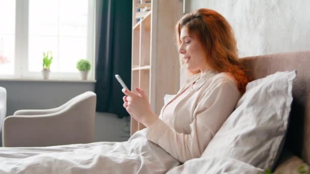 白种人女人在床上安安静静的早上例行公事醒来后睡小玩艺儿网瘾女性美女影响者浏览手机聊天社交媒体笑着 — 图库视频影像