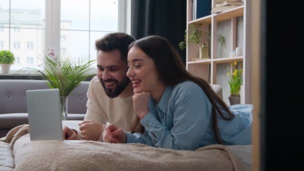 幸せな白人女性と男ベッドの上に横たわる屈託のないカップル見る面白いビデオでラップトップでベッドルーム笑顔夫のボーイフレンドと妻のガールフレンドホームオーナーサーフィンコンピュータウェブサイト楽しい喜び笑いを持っています — ストック動画