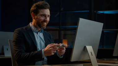 Adult businessman employee working in evening office happy male gamer Caucasian man take break playing console video game on computer with gaming controller using joystick gamepad play virtual match