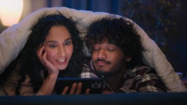 Çok ırklı çift ve kadın gece yorganın altında cep telefonuyla internette gezinerek akıllı telefon aile aleti bağımlısıyla video alışverişini tartışarak mutlu mesut kamuflaj sağlıyorlar.