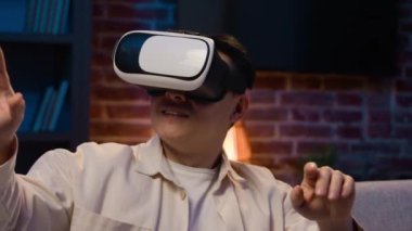 Asyalı adam video oyunu oynuyor sanal gerçeklik tecrübesi siber uzay modern teknoloji gece evde 3D gözlükler gözlüklü Koreli gözlüklü adam VR gözlüklü adam akşamları koltukta simülasyon oynuyor.