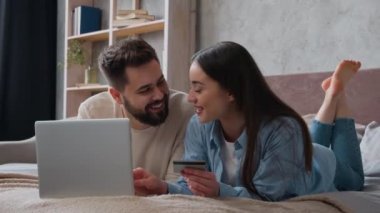 Kafkasyalı çift yatakta yan yana yatıyorlar kredi kartı kullanıyorlar dizüstü bilgisayar bankacılığı uygulamasına şifreyi giriyorlar gülümseyen kadın ve erkek alışveriş siparişleri ev yatak odasında ödeniyor.