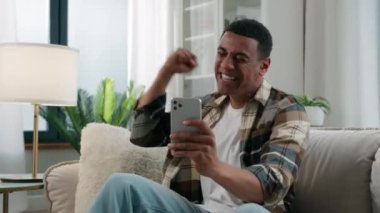 Mutlu Afrikalı Amerikalı kullanıcı kazananı cep telefonuyla zaferi kutluyor. Akıllı telefon galibiyeti. Kanepede cep telefonu ödüllü koltukta kazanmak.
