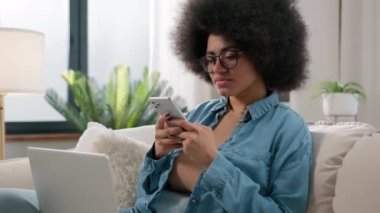 Kanepe üzerinde dizüstü bilgisayarı olan Afrikalı Amerikalı etnik kız serbest çalışan cep telefonuyla uzaktan kumandalı iş e-öğrenme bilgisayarına kayan akıllı telefon uygulaması kullanarak ara verdi.
