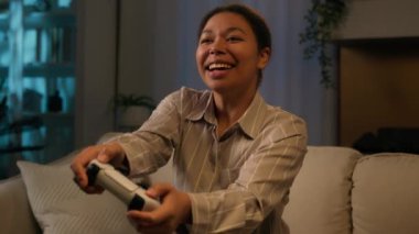 Afro-Amerikalı kadın oyuncu, evinde kanepede oturup oyun oynarken mutlu oluyor. Gülümseyen bir kadın, kumandalı sanal sanal uzay teknolojisiyle oyun oynuyor. Akıllı TV 'de oturma odasında.