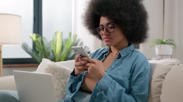 非洲裔美国女性自由撰稿人 带着笔记本电脑在沙发上浏览手机 在遥远的工作岗位上休息使用滚动智能手机应用程序远程学习电脑在家里阅读令人困惑的新闻 — 图库视频影像