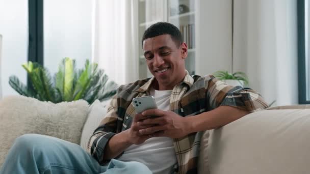 アフリカ系アメリカ人の男幸せな笑顔男は モバイルアプリケーションを使用して自宅で快適なソファソファの上でリラックス携帯電話アプリをスクロールチャットソーシャルネットワークオンラインサーフィンインターネットスマートフォンネット — ストック動画