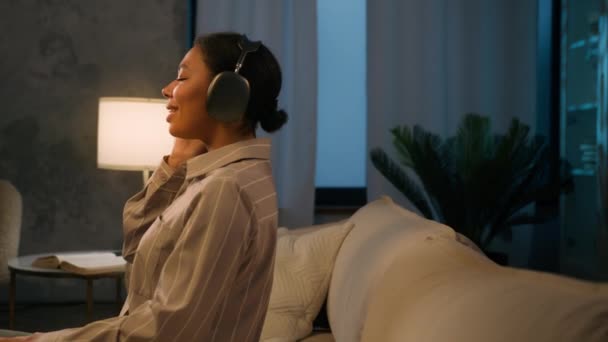 厌倦了的非洲裔美国女人晚上在沙发上放松 客厅里用无线耳机享受音乐 靠在舒适的沙发上休息 闭着眼睛闭着眼睛听着轻松的音乐唱歌 — 图库视频影像