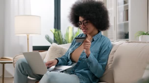 アフリカ系アメリカ人女性の顧客のソファインターネットショッピングでクレジットカードをオンラインで購入ウェブ支払い入力ノートパソコンで自宅の女の子の買い物客は ウェブサイト上で購入を支払う注文食品配達予約 — ストック動画