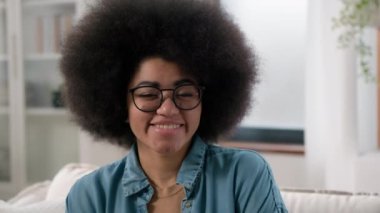 Neşeli yalnız genç Afrikalı Amerikalı kadın kanepeye gülümseyen portre gözlüklü mutlu etnik kız ev hanımı kız arkadaş kameraya bakıp kendi malikanesinde gülümsüyor.