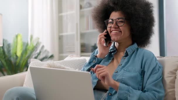アフリカ系アメリカ人の女の子がノートパソコンで作業して携帯電話を話すオンライン注文自宅でソファで食品配達を予約するビジネスコールを話すコンピュータプロジェクトについて話し合うフレンドリーな会話 — ストック動画