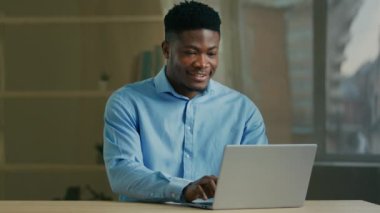 Gülümseyen Afrikalı Amerikalı işadamı ofiste bilgisayar kullanıyor. Erkek profesyonel yönetici dizüstü bilgisayarda yazarken gülümsüyor e-posta yazıp internette e-ticaret projesi üzerinde çalışıyor.