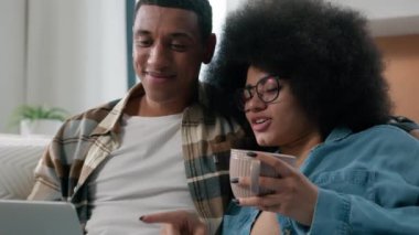 Mutlu aile çifti Afrikalı Amerikalı koca kanepeye uzanmış bir fincan çay içerken dizüstü bilgisayar alışverişi yapıyor internet mağazası uygulamasında ürünler seçiyor sipariş teslimatını birlikte tartışıyorlar.