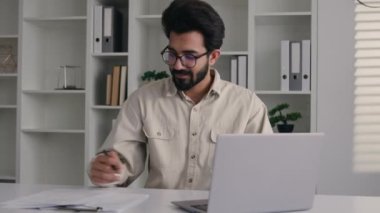 Arap müslüman girişimci erkek Hintli iş adamı dizüstü bilgisayar yazma projesinde çalışıyor bütçe planı yazıyor gelir finansmanı internet faturaları ofis evrakları kameraya gülümsüyor