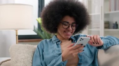 Afro-Amerikalı mutlu bir kadın kanepe kanepesinde dinleniyor. Mobil uygulama, kaydırmalı telefon uygulaması, sosyal ağla sohbet, internet sörfü, akıllı telefon alışveriş siparişi.