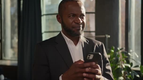 成熟した40アフリカ系アメリカ人のビジネスマンオフィスで携帯電話にメッセージを入力することを考えている携帯電話に間違いないと思うビジネスアイデア幸せな労働者が良い決定を思い付く — ストック動画