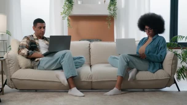 アフリカ系アメリカ人のカップル技術ビデオゲーム中毒家族と2台のラップトップはソファの上でオンラインでプレイホーム女性はソファの上でコンピュータでフリーランスと一緒に働いているラップトップの男と仕事は無視 — ストック動画