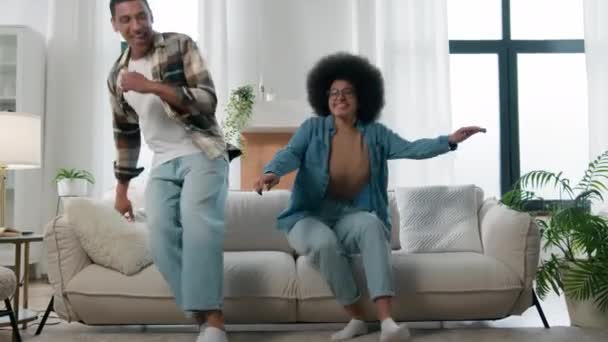 幸せなアフリカ系アメリカ人のカップルはソファの上でジャンプ笑いながら楽しい時間を過ごしてリラックスした休息をお楽しみください居心地の良いソファで秋を祝います移動日の移転を自分の家にレンタルフラット面白い家の所有者家族 — ストック動画