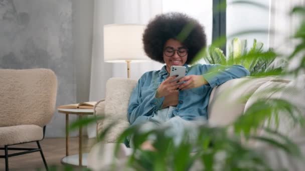家の植物を通して見る幸せな笑顔リラックスアフリカ系アメリカ人女性気楽な女の子休みます上のソファに自宅でスクロールソーシャルメディアネットワーク笑顔モバイルアプリケーションブラウジング電話チャットオンライン — ストック動画